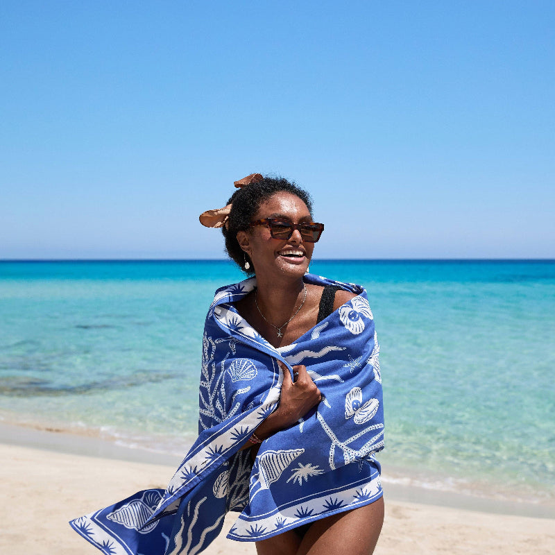 Woman wrapped in a blue and white patterned towel on a beach with clear blue water and sky.