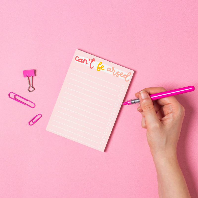 Hand holding a pink pen over a notepad with 'can't be arsed' text on a pink background