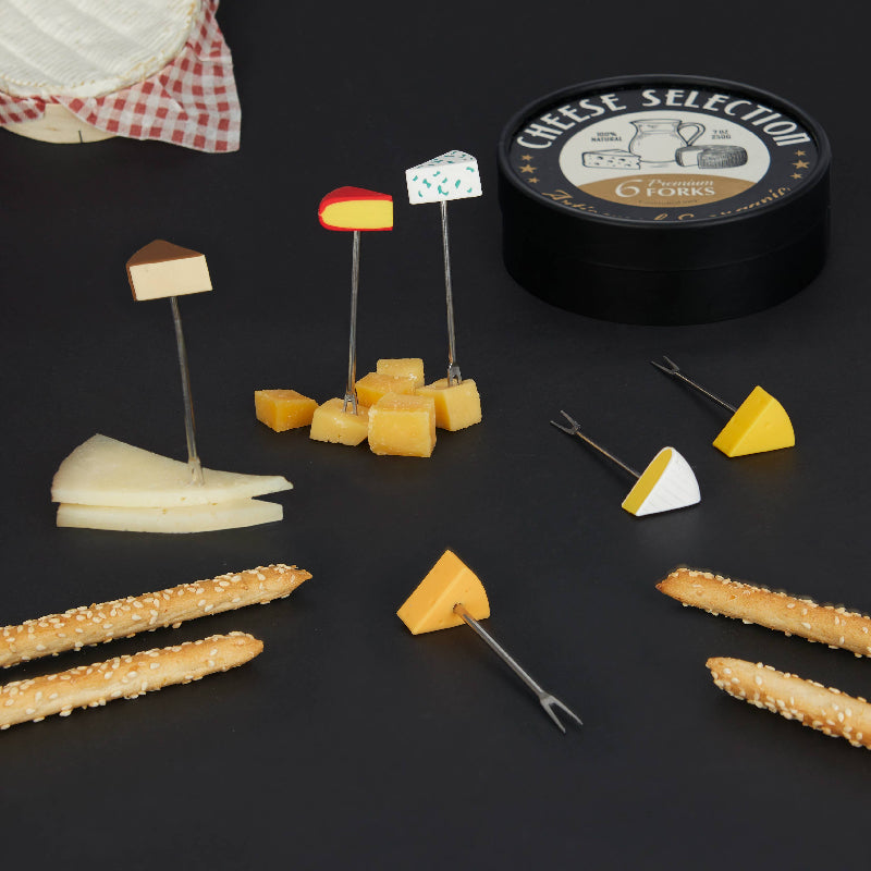 A cheese selection set displayed on a dark surface, including a round black tin labeled 'CHEESE SELECTION' and '6 FORKS', several cheese wedges and cubes on picks, two pretzel sticks, and two cheese forks.