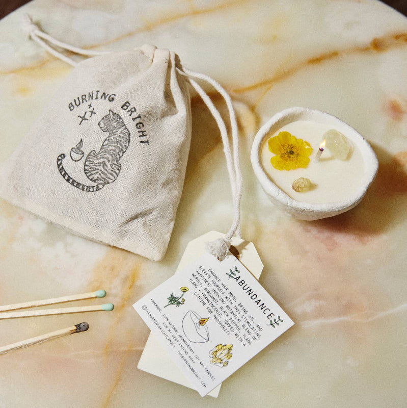 Candle with floral elements, matches, and a branded drawstring bag on a marble surface