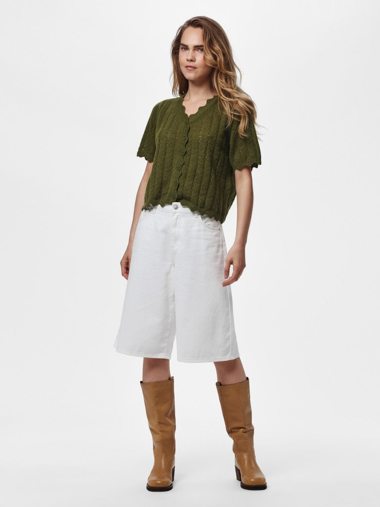 A woman modeling a short-sleeve olive green knit top with white wide-leg shorts and tan knee-high boots, photographed against a plain light gray studio background.