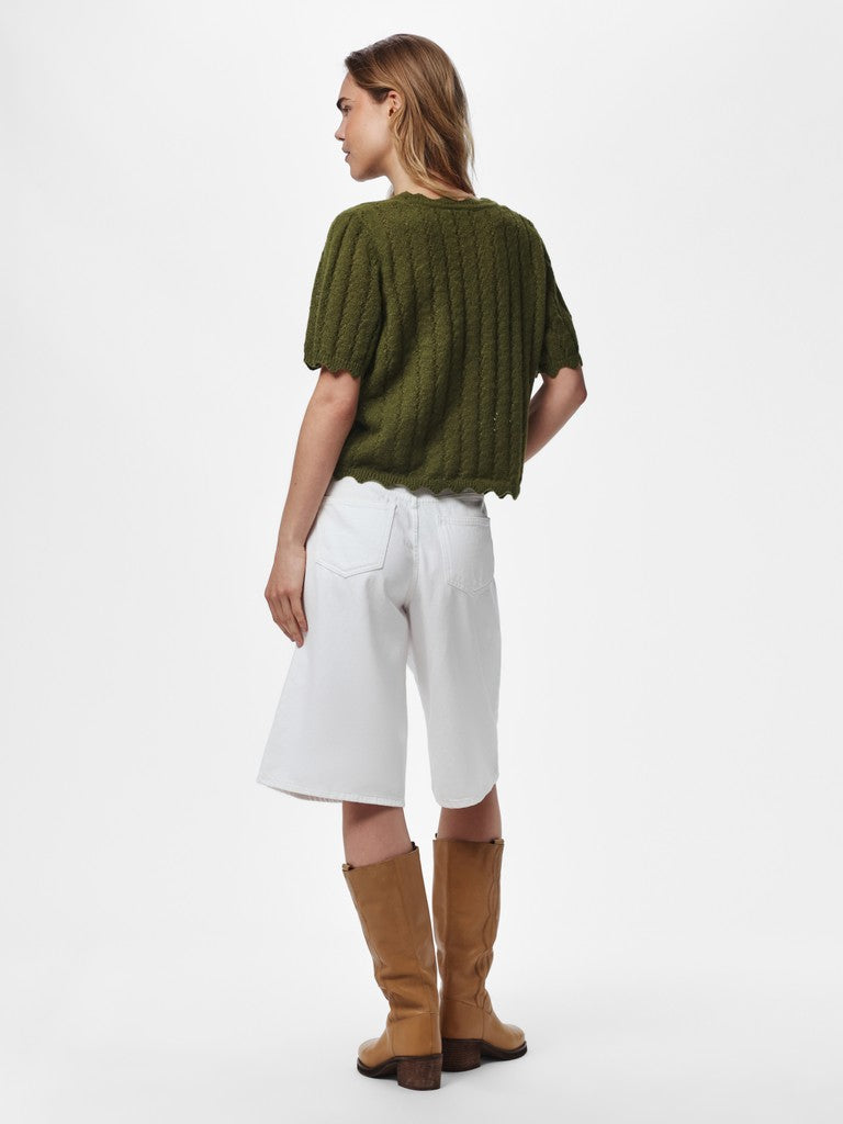 A woman shown from the back wearing an olive green short-sleeve knit top, white cropped shorts, and tan knee-high boots against a plain light gray studio background.