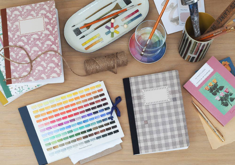 Tabletop with art supplies including notebooks, paintbrushes, and color swatches.
