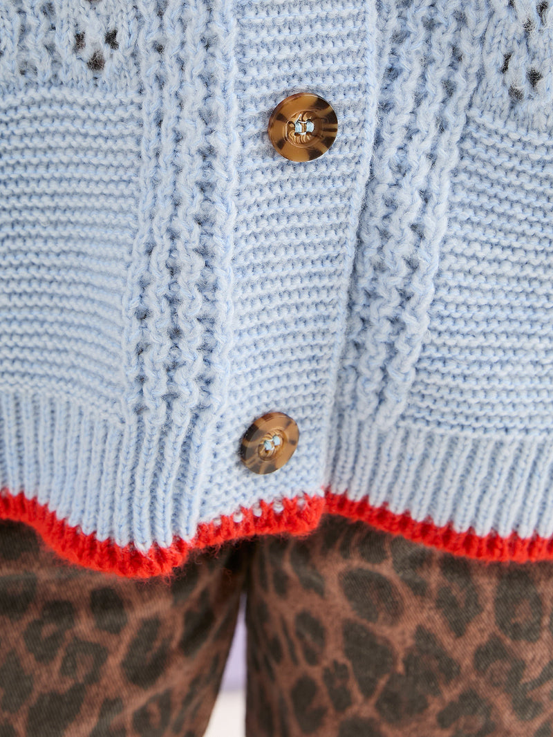 Close-up of a light blue knitted cardigan with brown buttons and red trim, worn over leopard print pants.