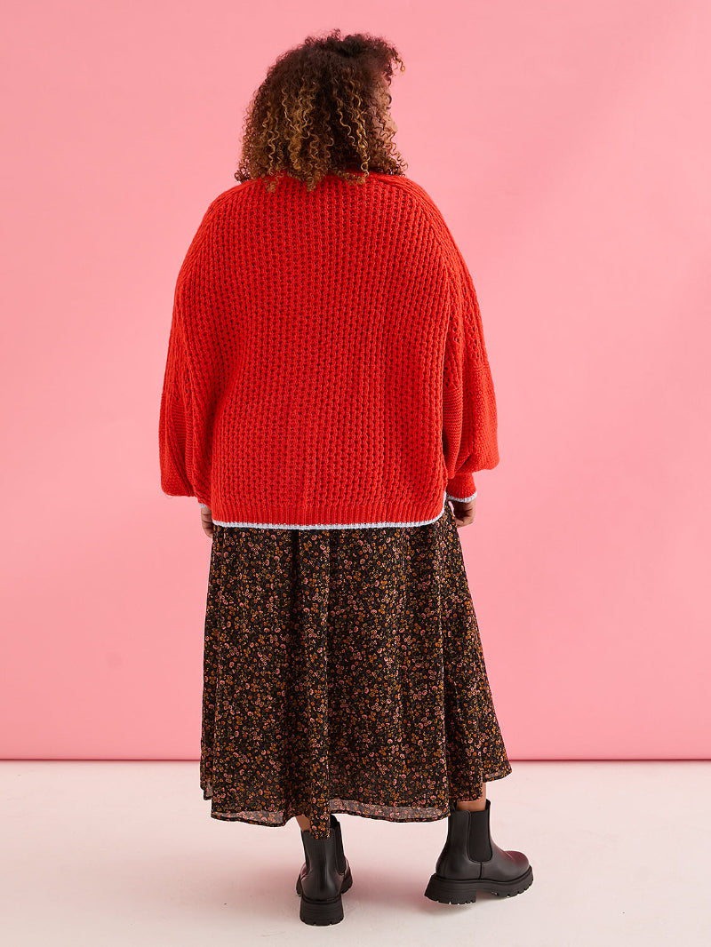 Person wearing Cara & The Sky Brigit Pointelle Cardigan a red sweater and patterned skirt against a pink background