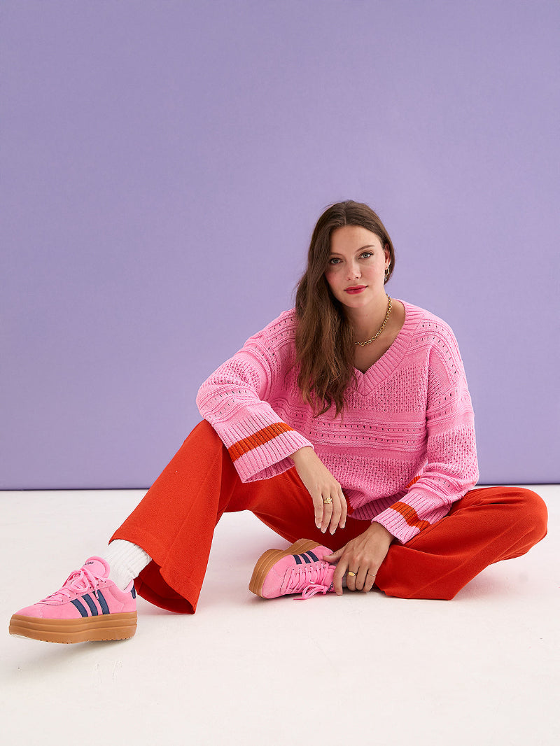 Woman wearing Cara & The Sky Judith Jumper a pink sweater and red pants sitting on a white floor with a purple background