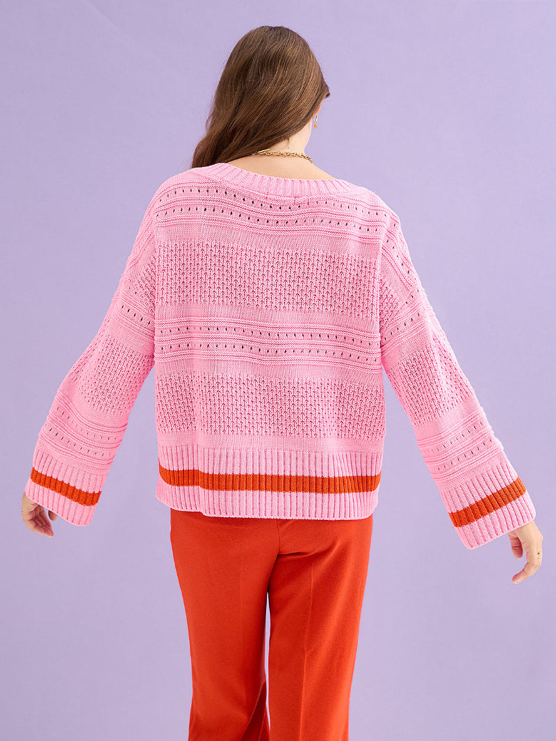 Person wearing Cara & The Sky Judith Jumper a pink sweater with a subtle pattern and red trim, standing against a light purple background.
