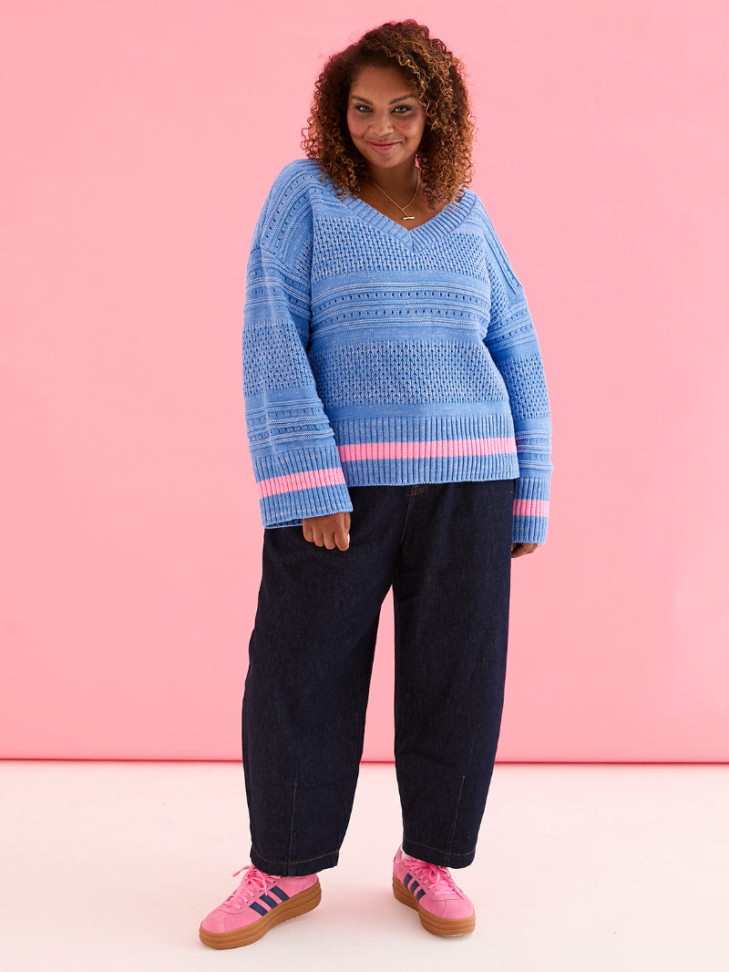 Woman wearing Cara & The sky JudithJumper Blue_Front a blue sweater with pink accents and pink sneakers against a pink background