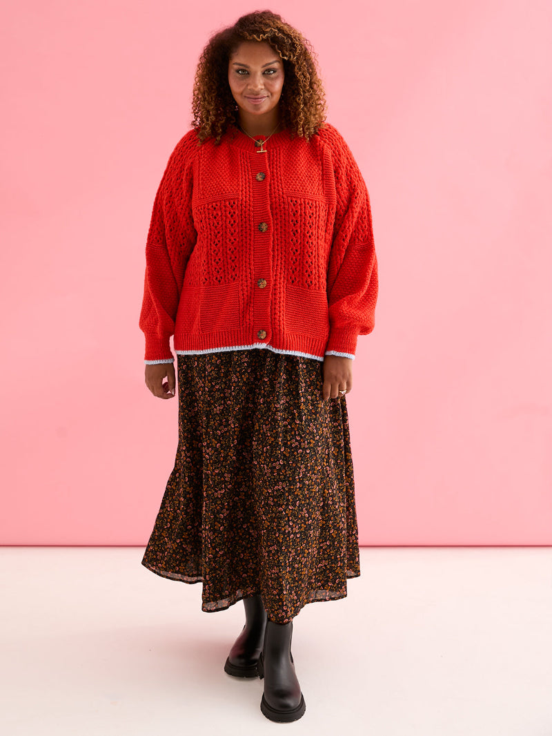 Woman wearing Cara & The Sky Brigit Pointelle Cardigan a red cardigan over a patterned dress against a pink background