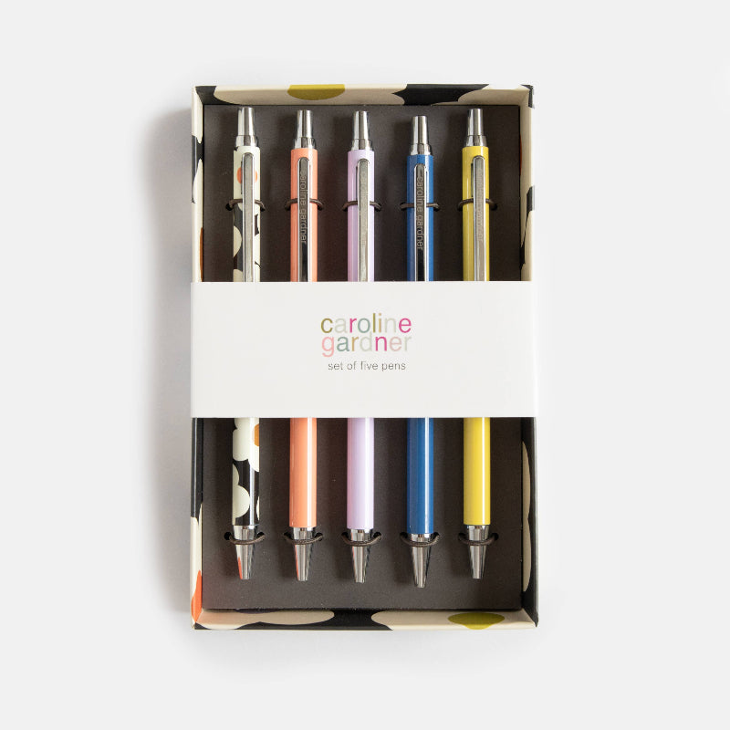 A set of five pens in a branded Caroline Gardiner gift box, arranged vertically with a white paper band across the middle. The pens are in silver, peach, lavender, blue, and yellow colors, set against a clean light gray background.