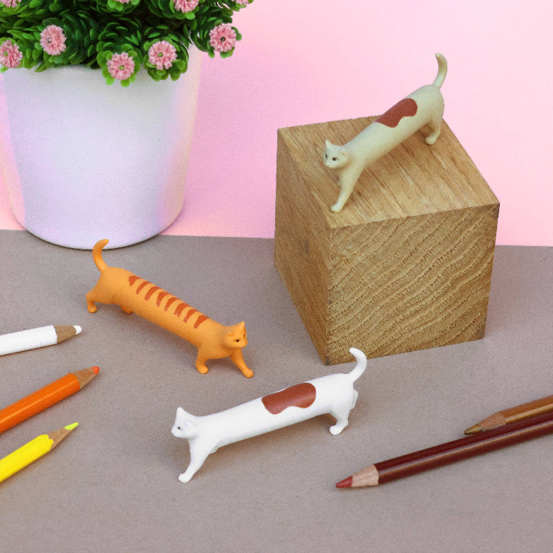 Cat-shaped pens on a desk with a wooden block and potted plant in the background