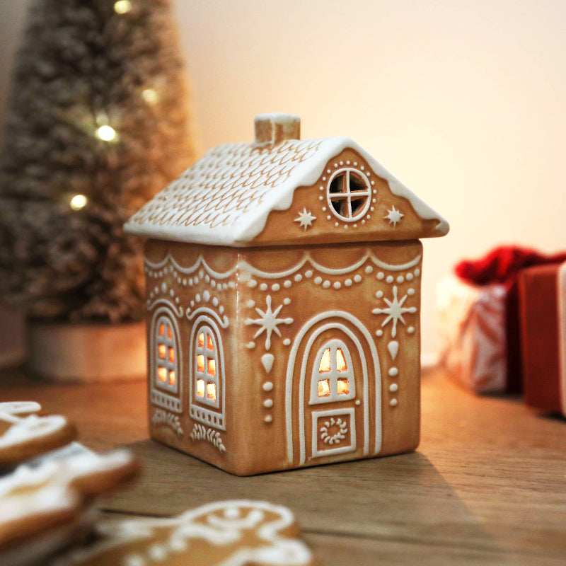 Decorative gingerbread house lantern on a wooden surface with blurred Christmas tree and gift in the background.