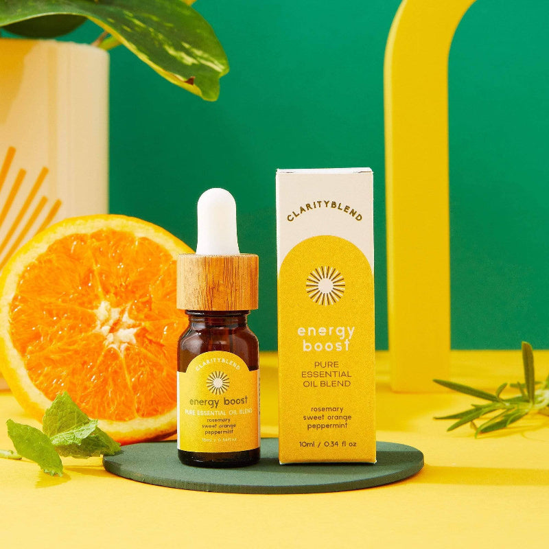 A small amber dropper bottle and its matching box for ClarityBlend Energy Boost pure essential oil blend, styled on a yellow surface with a green tray, a sliced orange, mint leaves, and a bright green and yellow background.