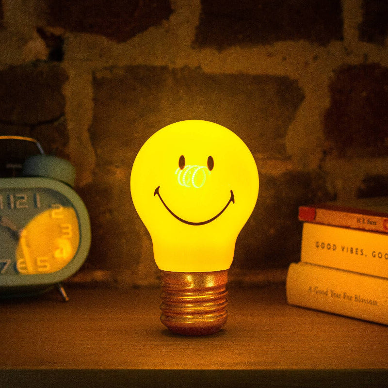 Smiley face light bulb on a wooden surface with books and an alarm clock in the background.