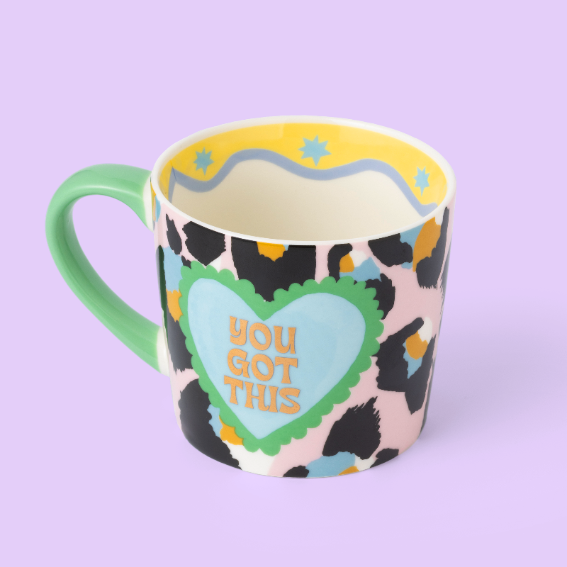 A ceramic mug with a multicolor leopard print pattern, a mint green handle, and a light blue heart graphic on the front that reads 'YOU GOT THIS' in orange. The mug is set against a solid lavender background.