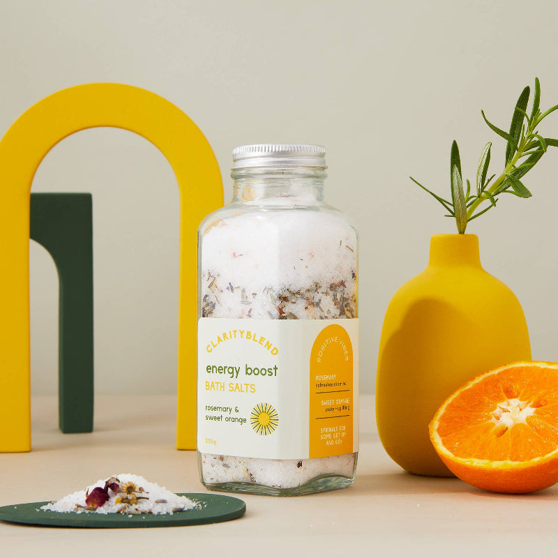 A glass jar of Charity Blend Energy Boost Bath Salts stands on a beige surface, styled with a yellow arch, a yellow vase with rosemary, and a halved orange. A small pile of the white bath salts with dried flower petals sits on a dark green plate in the foreground.