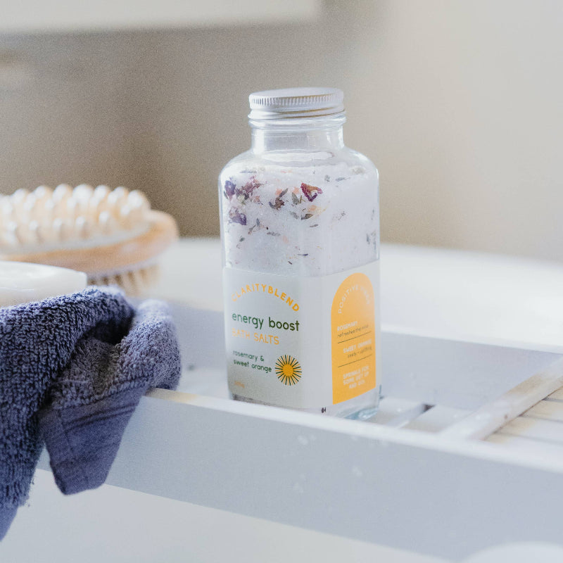 A clear glass bottle of bath salts labeled 'energy boost' with dried botanicals inside, set on a white bathtub ledge next to a blue towel and soap dish.