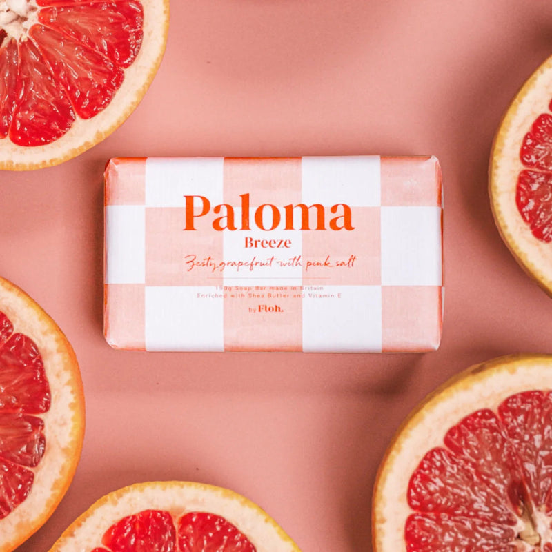 Bar of soap with 'Paloma Breeze' packaging surrounded by sliced grapefruits on a pink background