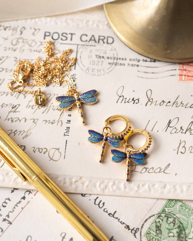 Gold and blue dragonfly-shaped earrings on a vintage postcard with a pen and gold object.