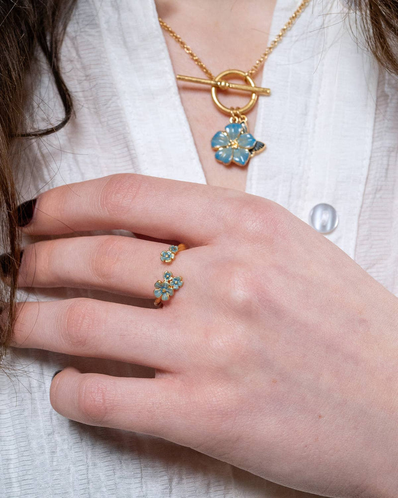 Close-up of a hand wearing a Fable Englande gold ring with blue flowers and a matching necklace with a blue flower pendant.