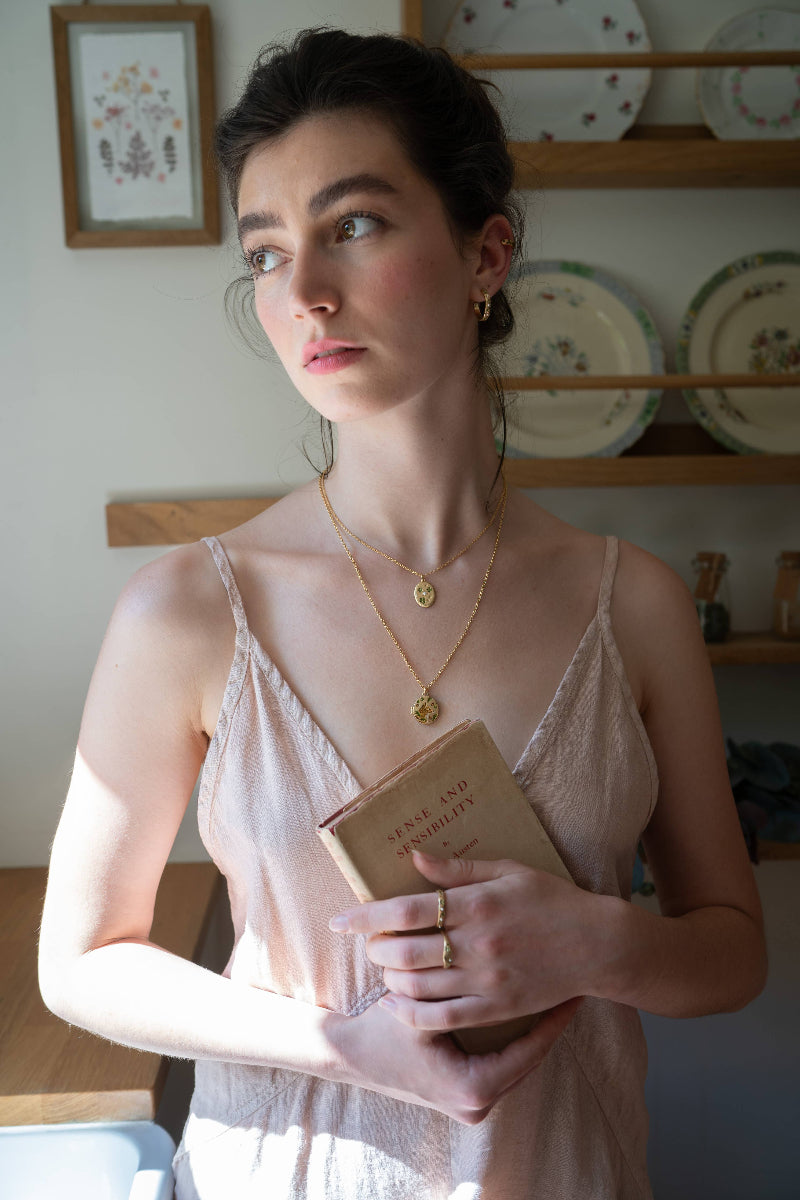 Woman holding a book in a kitchen setting with decorative plates on the wall.Wearing Fable Fox & Acorn Oval Chain Locket Necklace