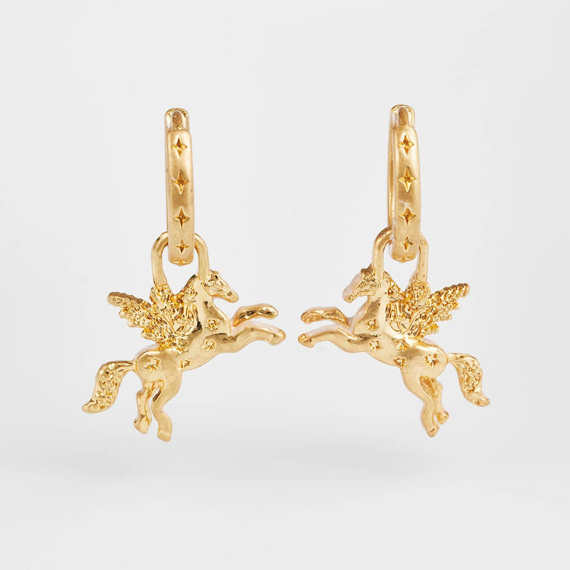 A pair of gold huggie hoop earrings featuring dangling winged horse figures, photographed against a clean light gray background.