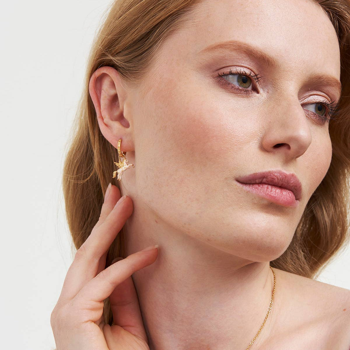 A close-up portrait of a woman with light brown hair wearing a delicate gold hoop earring with a small star detail, along with a fine gold chain necklace, against a clean white background.