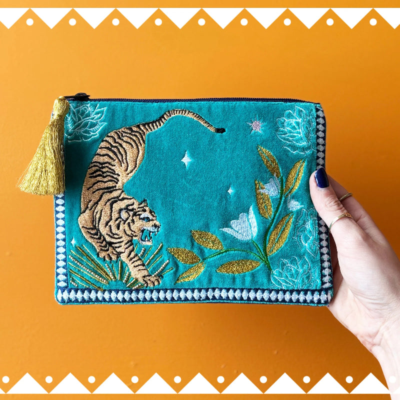 A turquoise embroidered pouch with a tiger, floral motifs, and a gold tassel, held by a hand against an orange background with a white geometric border.