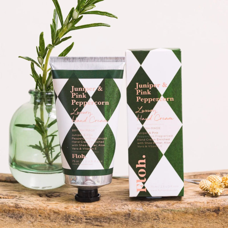 Floh. Juniper & Pink Peppercorn Lavender Hand Cream shown as a squeeze tube next to its retail box, styled on a rustic wooden surface with a green glass vase containing rosemary sprigs.