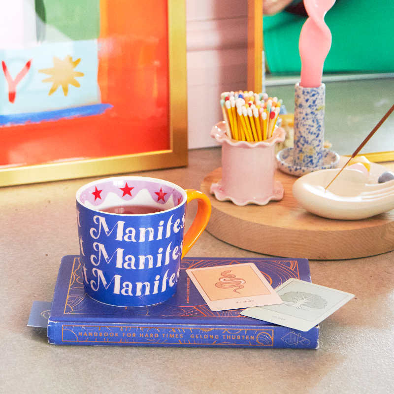 Blue mug with 'Manife' text on a table with books and cards