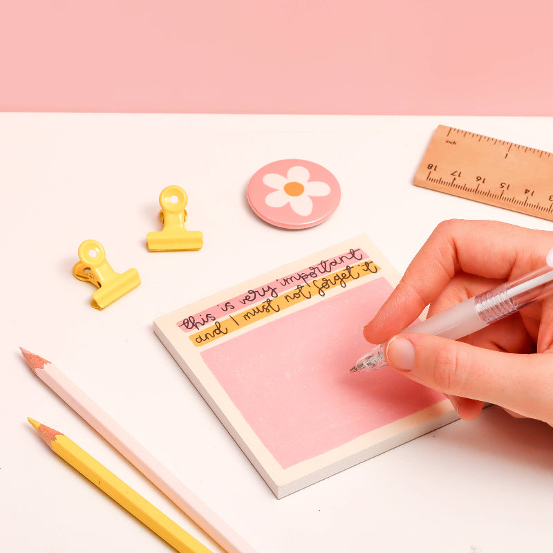 Person writing on a pink sticky note with stationery items on a light pink background