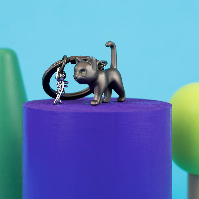 A small black cat figurine holding a silver keyring, displayed on top of a purple cylindrical pedestal against a bright blue background with blurred green shapes.