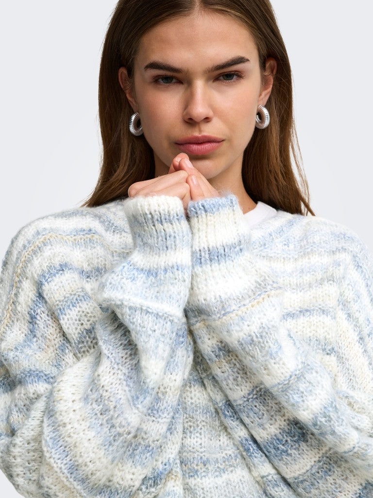 A close-up of a woman wearing a chunky knit sweater with marled light blue, white, and beige stripes. She is looking directly at the camera with her hands crossed under her chin, wearing silver hoop earrings against a plain light gray background.