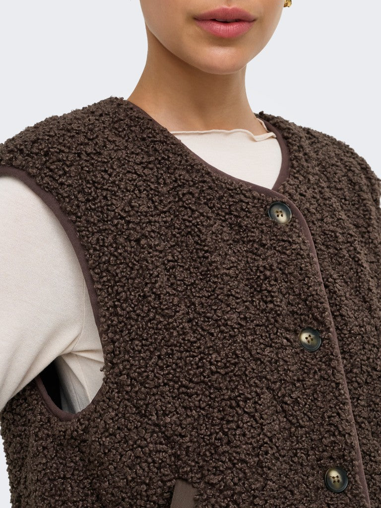 Close-up of a person wearing a brown textured fleece button-front vest over a cream top, against a light gray background.