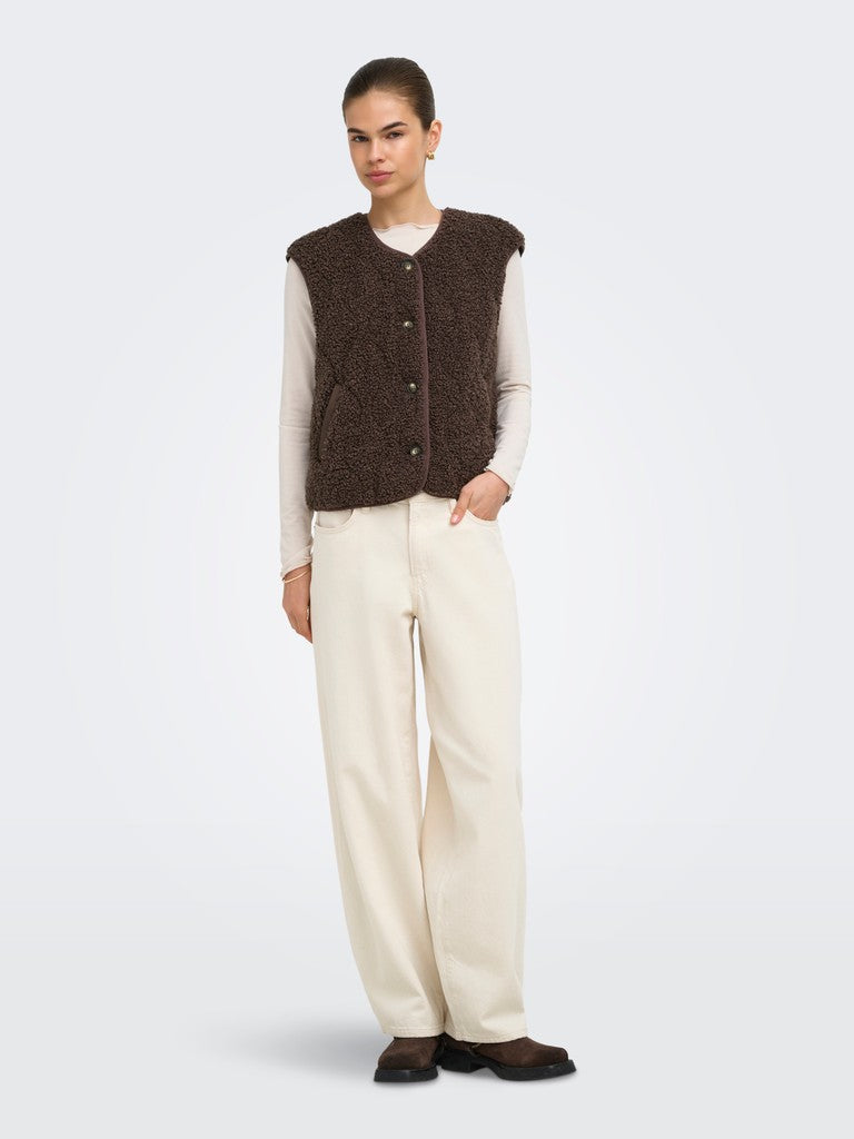 A model stands against a light gray studio background wearing a dark brown textured knit sleeveless vest over a cream long-sleeve top and cream wide-leg trousers.