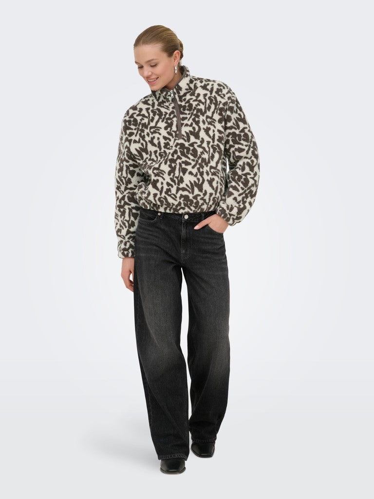 A woman modeling a black and beige animal print quarter-zip fleece jacket paired with black jeans, standing against a light gray studio background.