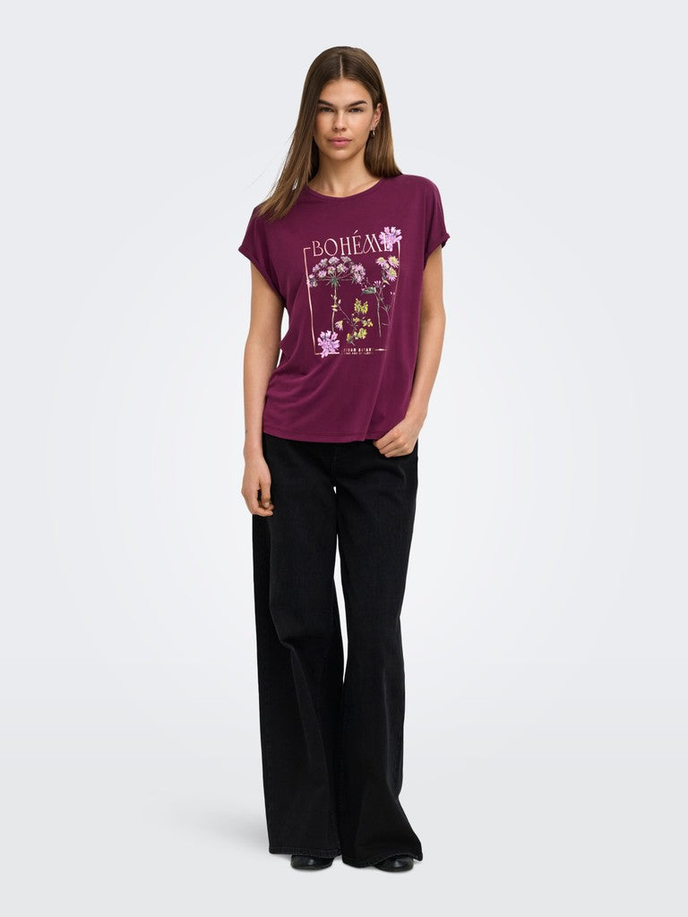 A woman modeling a short-sleeve crewneck t-shirt in deep purple with a pink and white floral graphic and the word 'BOHEM' on the front, paired with black wide-leg trousers, against a light gray studio background.