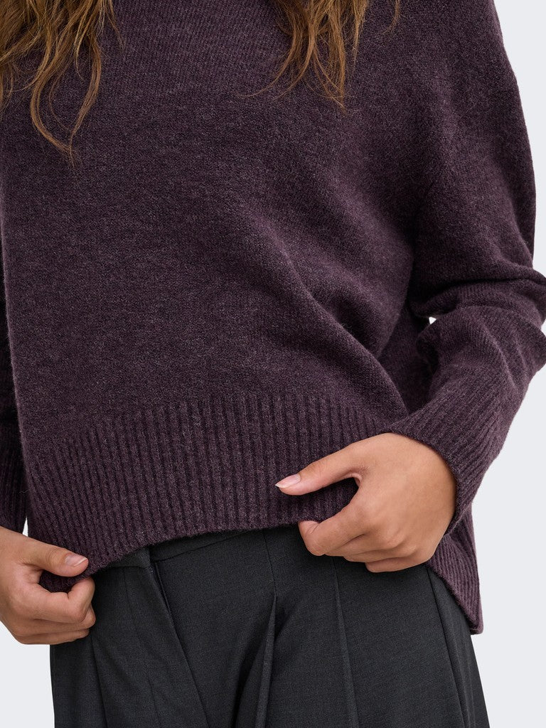 Close-up of a person wearing a dark plum ribbed knit sweater with black trousers, showing the texture and fit of the sweater.