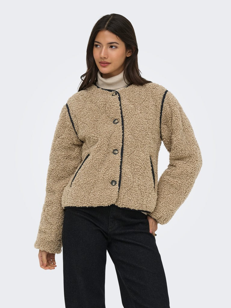 A woman modeling a cropped beige sherpa fleece jacket with black piping and button details, paired with black pants, against a light gray studio background.