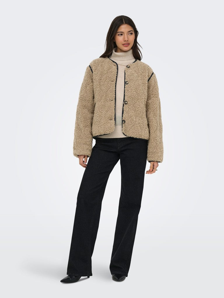 A model stands against a light gray studio background wearing a cropped, textured beige sherpa-style jacket with dark trim over a cream turtleneck and black wide-leg trousers.