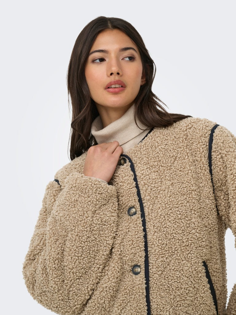 A woman wearing a beige sherpa fleece button-up jacket with dark contrast piping over a cream turtleneck, photographed against a light gray studio background.