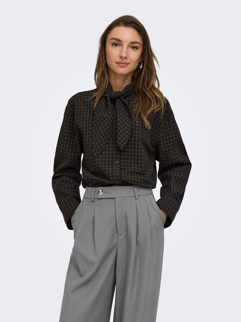 A woman modeling Only Maya Check Bow Shirt a dark brown and black checkered long-sleeve blouse with a tie-neck detail, paired with high-waisted gray trousers, against a light gray studio background.