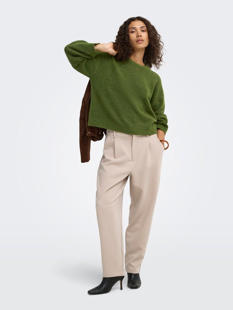 A woman modeling a green knit sweater, beige wide-leg trousers, and black ankle boots, with a brown jacket draped over her shoulder, against a light gray studio background.