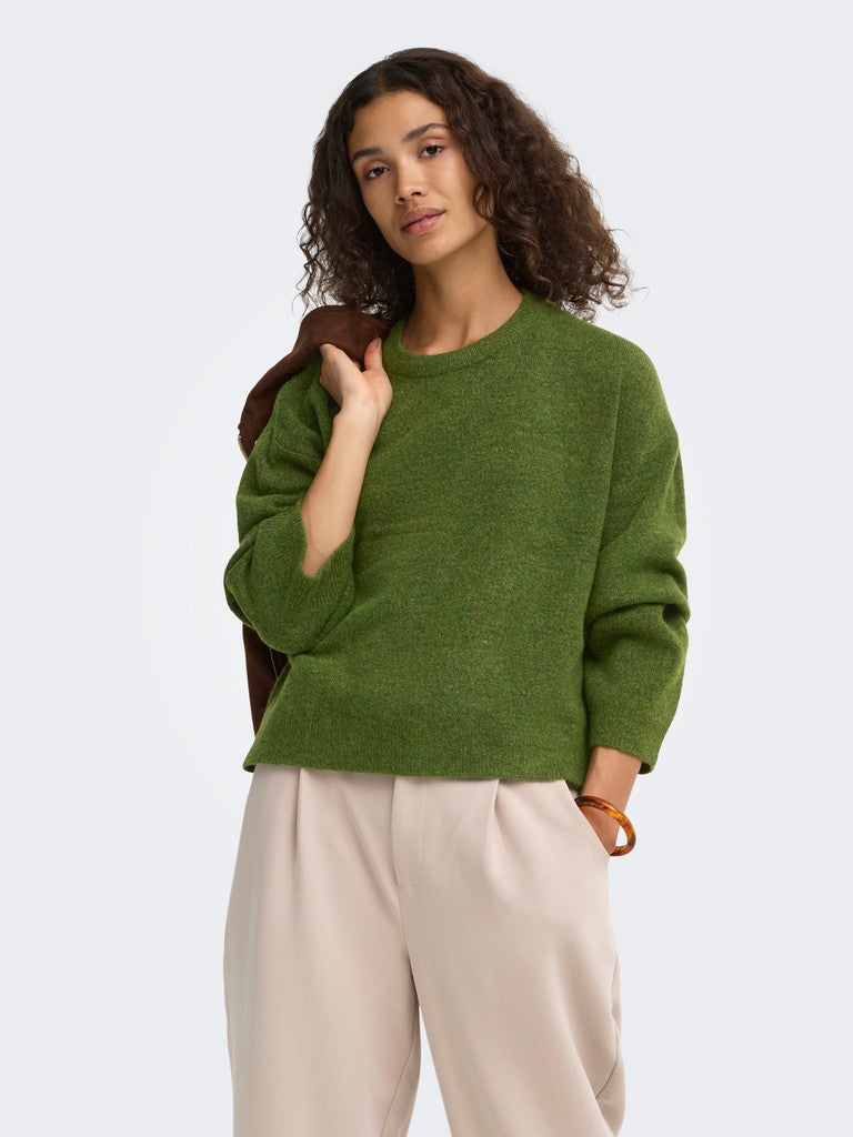 A woman modeling a green crewneck knit sweater and light beige trousers against a plain light gray studio background.