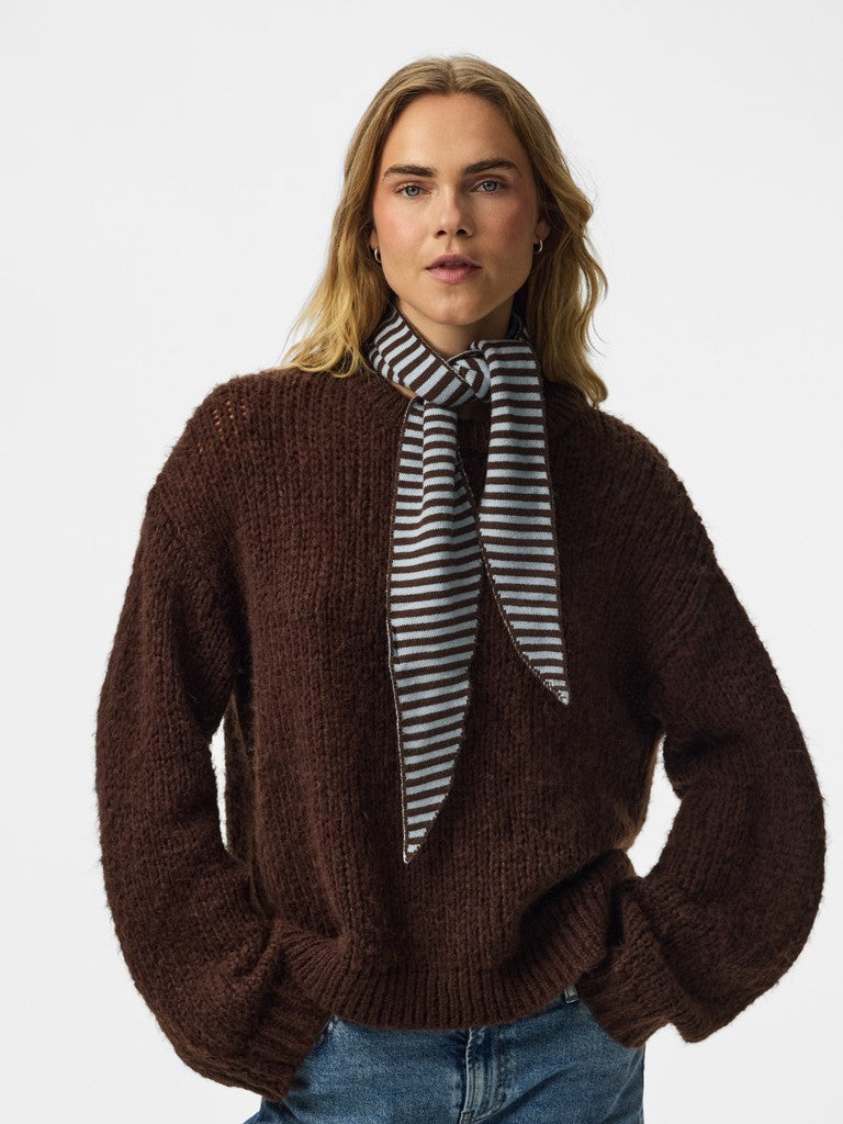 A woman wearing a chunky dark brown knit sweater, a blue and brown striped neck scarf, and blue jeans, photographed against a plain light gray studio background.