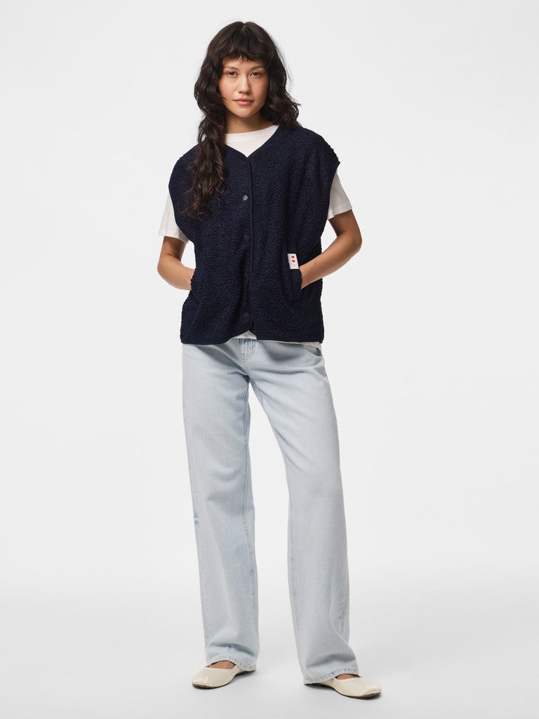 A model stands against a light gray studio background wearing a dark navy sleeveless button-front vest over a white t-shirt, paired with light blue straight-leg jeans and beige shoes.