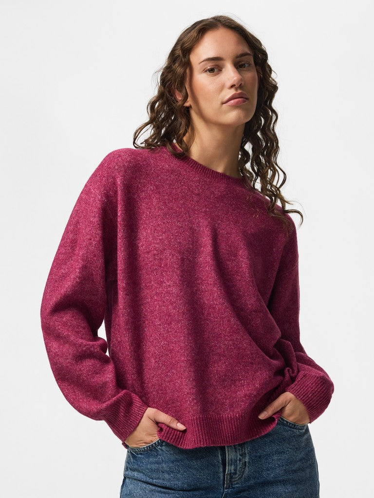 A woman wearing a heathered magenta knit sweater and blue jeans, standing against a plain white studio background.