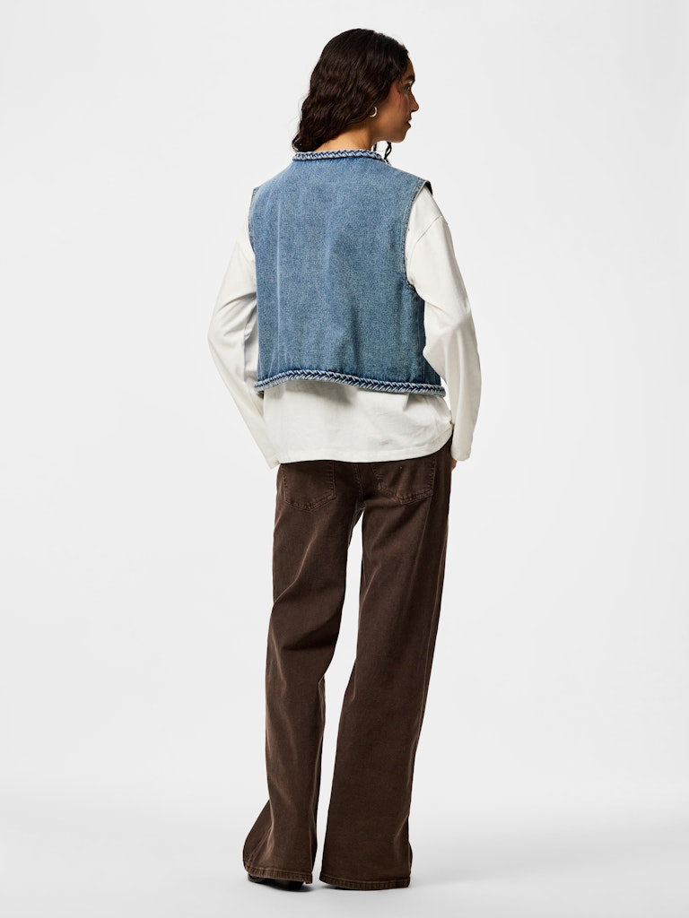 Person wearing a denim vest over a white shirt with brown pants on a white background