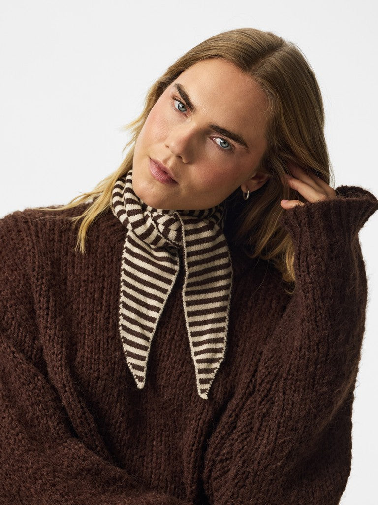 A woman wearing a chunky dark brown knit sweater and a brown and cream striped scarf, photographed against a clean light gray studio background.