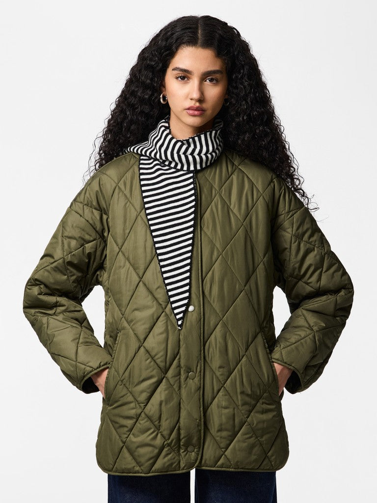 A model with long, curly dark hair is wearing an olive green diamond-quilted jacket over a black and white striped shirt, paired with dark blue jeans, against a plain light gray studio background.